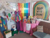 Raglan Area School Out on the Shelves display 2022. Rainbows decorate a table with books displayed on it. A mannequin wears a rainbow tulle skirt. There is a rainbow flag and a sign saying "Read with pride" behind the table of books and a couch.